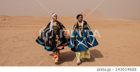 Two male Egyptian tanoura folk performers in contrasting black and teal embroidered robes and turbans walking across Giza desert plateau with ancient Two male Egyptian tanoura folk performers in contrasting black and teal embroidered robes and turbans walking across Giza desert plateau with ancient 143014688