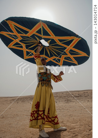 Egyptian tanoura dancer in yellow embroidered robe raises enormous teal geometric spinning disc overhead in open sandy desert under blazing midday sun Egyptian tanoura dancer in yellow embroidered robe raises enormous teal geometric spinning disc overhead in open sandy desert under blazing midday sun 143014714