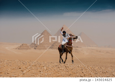 Exhilarated Arab horseman on galloping brown stallion raises arm before sweeping panorama of all three Giza pyramids on rocky sandy desert plateau Exhilarated Arab horseman on galloping brown stallion raises arm before sweeping panorama of all three Giza pyramids on rocky sandy desert plateau 143014825