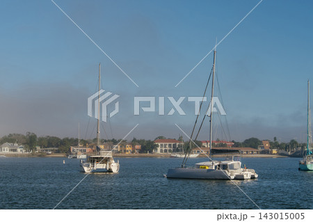 Two large catamarans float on blue water near the Saint Augustine shoreline. Colorful waterfront homes and low morning fog appear behind the anchored boats. 143015005