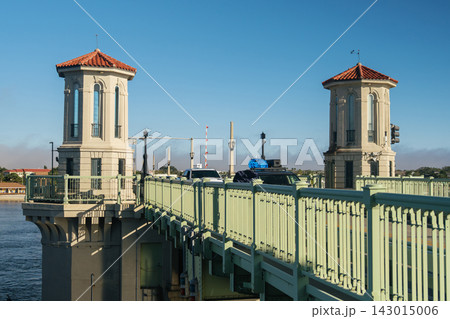 Two Mediterranean-style towers rise above the Bridge of Lions in Saint Augustine, Florida. Cars cross the green drawbridge beside blue water under a clear sky. 143015006
