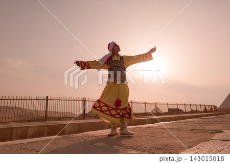 Whirling dervish style performer in yellow embroidered Egyptian folk costume arms outstretched against a warm hazy golden sunset sky at the Giza Whirling dervish style performer in yellow embroidered Egyptian folk costume arms outstretched against a warm hazy golden sunset sky at the Giza 143015018