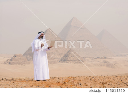Ambitious Arab entrepreneur in traditional white kandura and ghutra using a laptop confidently outdoors on the sandy Giza plateau with the iconic Ambitious Arab entrepreneur in traditional white kandura and ghutra using a laptop confidently outdoors on the sandy Giza plateau with the iconic 143015220