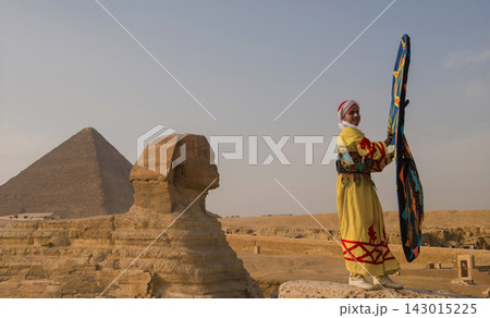 Colorful Egyptian folk performer in embroidered yellow robe holds decorative shield standing beside Great Sphinx of Giza with ancient pyramid in sandy Colorful Egyptian folk performer in embroidered yellow robe holds decorative shield standing beside Great Sphinx of Giza with ancient pyramid in sandy 143015225