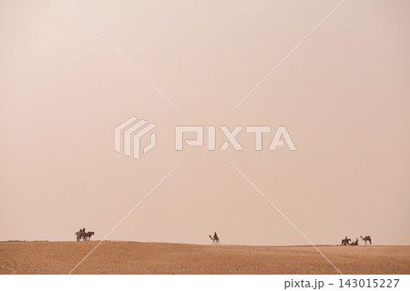 Camel riders and horse cart traverse a sweeping Egyptian desert plateau under enormous hazy pale sky ideal for MENA travel editorial and minimalist 143015227