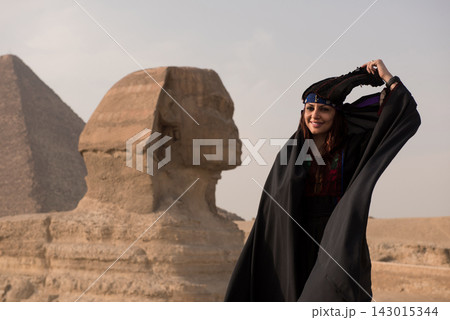 Joyful young Middle Eastern woman in ornate embroidered black folk costume playfully adjusting beaded head wrap with the iconic Great Sphinx and Giza Joyful young Middle Eastern woman in ornate embroidered black folk costume playfully adjusting beaded head wrap with the iconic Great Sphinx and Giza 143015344