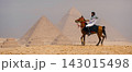 Bearded Arab horseman in white shirt and keffiyeh rides a chestnut horse mid stride on the rocky Giza plateau with two ancient pyramids rising through 143015498
