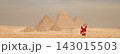 Playful Santa Claus figure in red suit waving on the sandy Giza desert plateau with the iconic Great Pyramids towering in the background under a hazy 143015503
