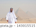 Cheerful Arab man wearing traditional white kandura and keffiyeh walking toward camera with soft focus Giza pyramids behind him evoking pride cultural 143015515