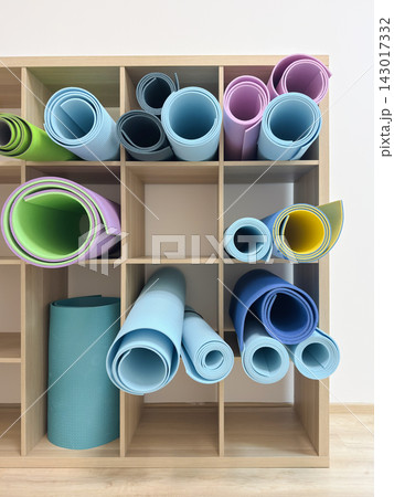 Rolled yoga mats fill a wooden storage shelf. Colorful fitness equipment is organized in compartments against a bright indoor wall. Active lifestyle and home workout organization Rolled yoga mats fill a wooden storage shelf. Colorful fitness equipment is organized in compartments against a bright indoor wall. Active lifestyle and home workout organization 143017332