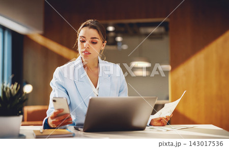 Busy businesswoman checks her smartphone while holding a printed report at her laptop in a warm wood paneled office Busy businesswoman checks her smartphone while holding a printed report at her laptop in a warm wood paneled office 143017536