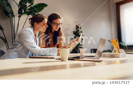 Creative business partners study a printed diagram over coffee at a sunlit table surrounded by lush green office plants 143017537