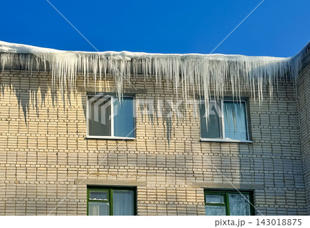 Long, powerful icicles hang from the snow-covered edge of the roof of a brick residential building. Long, powerful icicles hang from the snow-covered edge of the roof of a brick residential building. 143018875