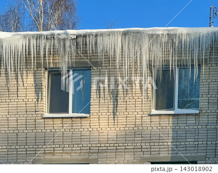 Long, powerful icicles hang from the snow-covered edge of the roof of a brick residential building. Long, powerful icicles hang from the snow-covered edge of the roof of a brick residential building. 143018902
