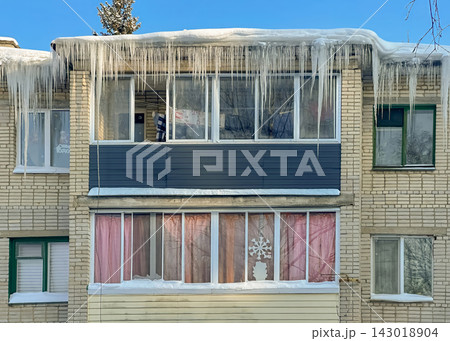 Long, powerful icicles hang from the snow-covered edge of the roof of a brick residential building. Long, powerful icicles hang from the snow-covered edge of the roof of a brick residential building. 143018904
