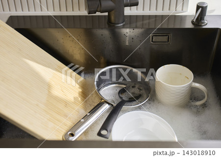 Saucepan, mug, bowl and cutting board soak in soapy water inside kitchen sink Saucepan, mug, bowl and cutting board soak in soapy water inside kitchen sink 143018910