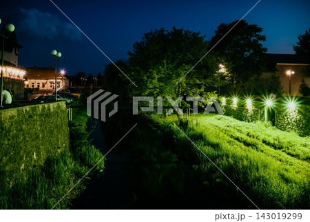 Beautiful night embankment near the river with green lights on a summer night. Weeping willow over the river at night. Green grass near the river at night. Beregovo. Ukraine Beautiful night embankment near the river with green lights on a summer night. Weeping willow over the river at night. Green grass near the river at night. Beregovo. Ukraine 143019299