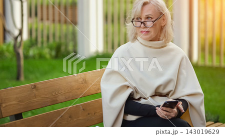 Older woman uses smartphone on park bench during quiet pause. Blonde woman sits in outdoor garden, wearing glasses and pale wrap, and studies her phone with thoughtful, focused expression. Older woman uses smartphone on park bench during quiet pause. Blonde woman sits in outdoor garden, wearing glasses and pale wrap, and studies her phone with thoughtful, focused expression. 143019629