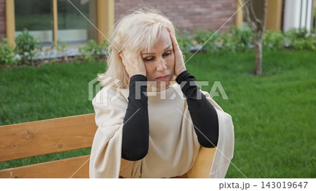 Woman presses both hands against ears while seated outdoors on wooden bench. Mature woman shows anxiety and sensory discomfort, surrounded by green grass, plants and residential windows. Woman presses both hands against ears while seated outdoors on wooden bench. Mature woman shows anxiety and sensory discomfort, surrounded by green grass, plants and residential windows. 143019647