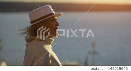 Woman in white hat looks toward tranquil water and glowing sunset. Older woman wears pale coat and scarf, her serene profile expressing quiet reflection beside softly blurred horizon. 143019648