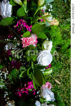 Sunlit floral arrangement with white roses, pink carnations, glossy green leaves, delicate filler blossoms, and dense textured foliage in vertical close-up composition for elegant decorative use 143019827