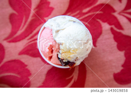 Overhead view of two scoops of pink and white ice cream in small bowl on vivid red floral fabric with soft blur, creamy texture, and playful summer dessert appeal Overhead view of two scoops of pink and white ice cream in small bowl on vivid red floral fabric with soft blur, creamy texture, and playful summer dessert appeal 143019858