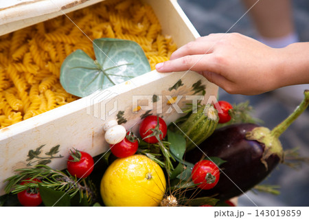 Child hand opening decorative box filled with fusilli pasta and fresh garden vegetables, with tomatoes, zucchini, eggplant, lemon, herbs, and leaves in colorful food arrangement 143019859