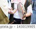 Close view of two people in traditional clothing with one holding frame drum and gesturing during outdoor cultural performance on city street with soft daylight and blurred background 143019905