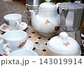 White floral teapot and matching cups with saucers beside metal coffee makers on mosaic tabletop in soft daylight, creating cozy tableware still life with mixed ceramic and steel textures 143019914