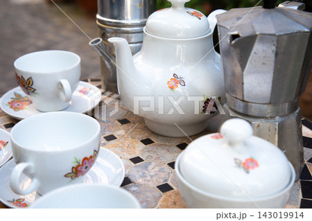 White floral teapot and matching cups with saucers beside metal coffee makers on mosaic tabletop in soft daylight, creating cozy tableware still life with mixed ceramic and steel textures White floral teapot and matching cups with saucers beside metal coffee makers on mosaic tabletop in soft daylight, creating cozy tableware still life with mixed ceramic and steel textures 143019914