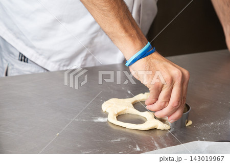 Close up of hand cutting rolled dough with round metal cutter on stainless steel counter in bright kitchen, showing pastry preparation, baking work, simple ingredients, and focused manual food craft 143019967