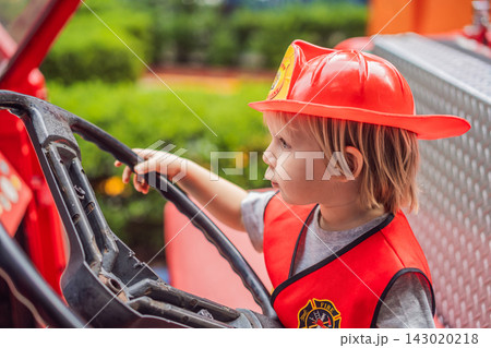 Little boy dressed as firefighter playing in a red toy fire truck at an outdoor playground with helmet safety vest pretend rescue game childhood dreams role play and wide view. 143020218