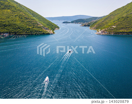 Aerial view of motorboats crossing the blue waters of the Bay of Kotor in Montenegro, surrounded by steep green coastal mountains near Lepetane and Kamenari. Aerial view of motorboats crossing the blue waters of the Bay of Kotor in Montenegro, surrounded by steep green coastal mountains near Lepetane and Kamenari. 143020879