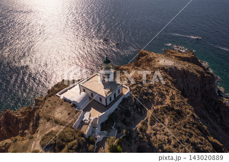 Elevated aerial view of Akrotiri Lighthouse on the rugged southwestern coast of Santorini, Greece, surrounded by shimmering Aegean Sea and rocky shoreline. Elevated aerial view of Akrotiri Lighthouse on the rugged southwestern coast of Santorini, Greece, surrounded by shimmering Aegean Sea and rocky shoreline. 143020889