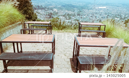 High-angle shot of an outdoor cafe seating area featuring wooden picnic tables and benches on a hillside terrace, overlooking a panoramic scenic view of a misty valley or mountain village, perfectly c High-angle shot of an outdoor cafe seating area featuring wooden picnic tables and benches on a hillside terrace, overlooking a panoramic scenic view of a misty valley or mountain village, perfectly c 143025005
