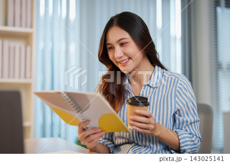 Smiling Asian woman holding coffee cup and reading notebook at office desk Smiling Asian woman holding coffee cup and reading notebook at office desk 143025141