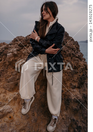 Woman leather jacket coastal rock sitting on cliff overlooking sea during overcast day for lifestyle fashion concept 143028275