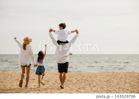 Amidst the sunset glow a happy family stands on the sea beach with raised arms. The father carrying his son on shoulders signifies the freedom and joy of a memorable beach vacation. Amidst the sunset glow a happy family stands on the sea beach with raised arms. The father carrying his son on shoulders signifies the freedom and joy of a memorable beach vacation. 143029786