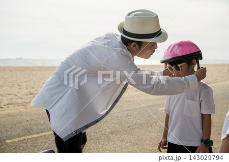 A family on vacation at the beach where the father sporting a safety helmet teaches his cheerful son the balance and joy of bicycle riding a memorable tourism day filled with family happiness. 143029794