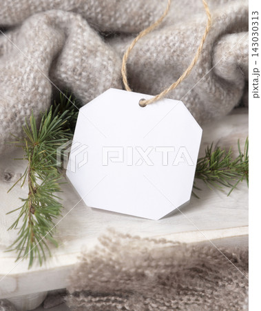 Blank gift tag mockup near beige knitted blanket and fir branches. Rustic Christmas packaging 143030313
