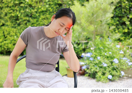 Unhappy Asian woman sitting on chair touching her head with red highlighted area indicating severe headache, migraine or brain pain. Concept of stress, sickness, healthcare, tension and mental health. 143030485