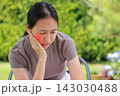 Unhappy young Asian woman sitting outdoors touching her cheek with a red highlighted area indicating severe toothache, dental pain, gum disease, or jaw problem. Healthcare, dentist, medical concept. 143030488