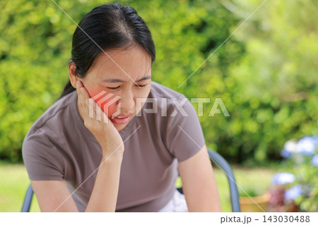 Unhappy young Asian woman sitting outdoors touching her cheek with a red highlighted area indicating severe toothache, dental pain, gum disease, or jaw problem. Healthcare, dentist, medical concept. Unhappy young Asian woman sitting outdoors touching her cheek with a red highlighted area indicating severe toothache, dental pain, gum disease, or jaw problem. Healthcare, dentist, medical concept. 143030488