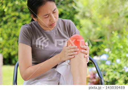Stressed young Asian woman sitting on a chair touching her bare knee with red highlighted area indicating severe joint pain, arthritis, muscle ache or sports injury. Healthcare, medical concept. 143030489