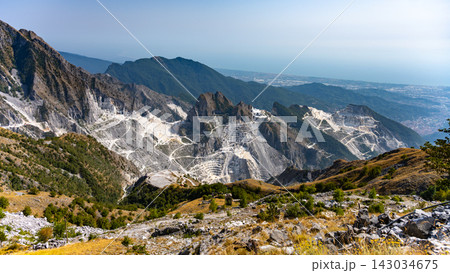 The image shows the Carrara Marble Quarries in Italy. White marble extraction areas are visible along with rocky hills and distant mountains. The scene highlights the mining activity and landscape. The image shows the Carrara Marble Quarries in Italy. White marble extraction areas are visible along with rocky hills and distant mountains. The scene highlights the mining activity and landscape. 143034675