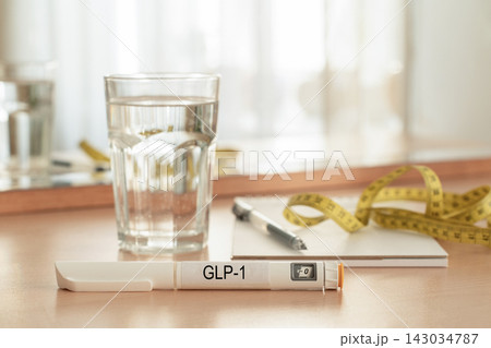 GLP-1 injection pen, measuring tape, notebook, and glass of water on wooden nightstand. Concept of medical weight loss, body weight control, health tracking, hydration, and healthy lifestyle GLP-1 injection pen, measuring tape, notebook, and glass of water on wooden nightstand. Concept of medical weight loss, body weight control, health tracking, hydration, and healthy lifestyle 143034787