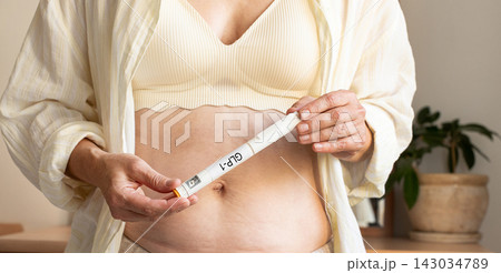 Woman holds GLP-1 pen injector against her abdomen at home. Concept of medical weight loss, obesity treatment, use of injectable medications, and body weight management Woman holds GLP-1 pen injector against her abdomen at home. Concept of medical weight loss, obesity treatment, use of injectable medications, and body weight management 143034789