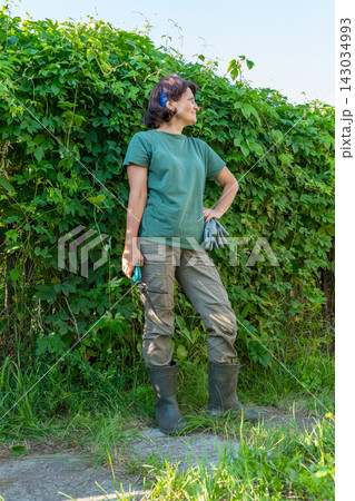 Female gardener in green attire standing by lush garden hedge outdoors Female gardener in green attire standing by lush garden hedge outdoors 143034993
