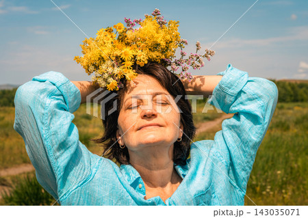 Woman with closed eyes and wreath of wildflowers on her head, wearing turquoise linen shirt, shot low against blue sky with light clouds. Women's mental health, slow living Woman with closed eyes and wreath of wildflowers on her head, wearing turquoise linen shirt, shot low against blue sky with light clouds. Women's mental health, slow living 143035071