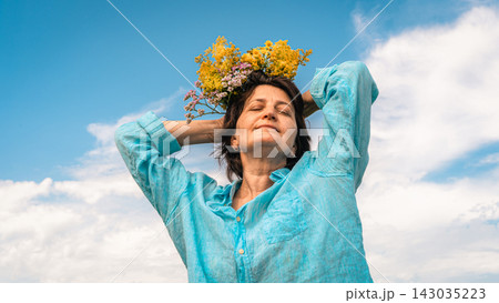 Woman with closed eyes and wreath of wildflowers on her head, wearing turquoise linen shirt, shot low against blue sky with light clouds. Women's mental health, slow living Woman with closed eyes and wreath of wildflowers on her head, wearing turquoise linen shirt, shot low against blue sky with light clouds. Women's mental health, slow living 143035223